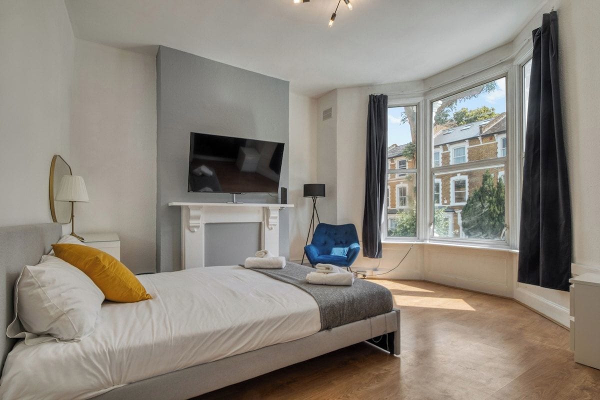 A bedroom with a bed, a flat-screen TV mounted above it, a chair, and a window with curtains.