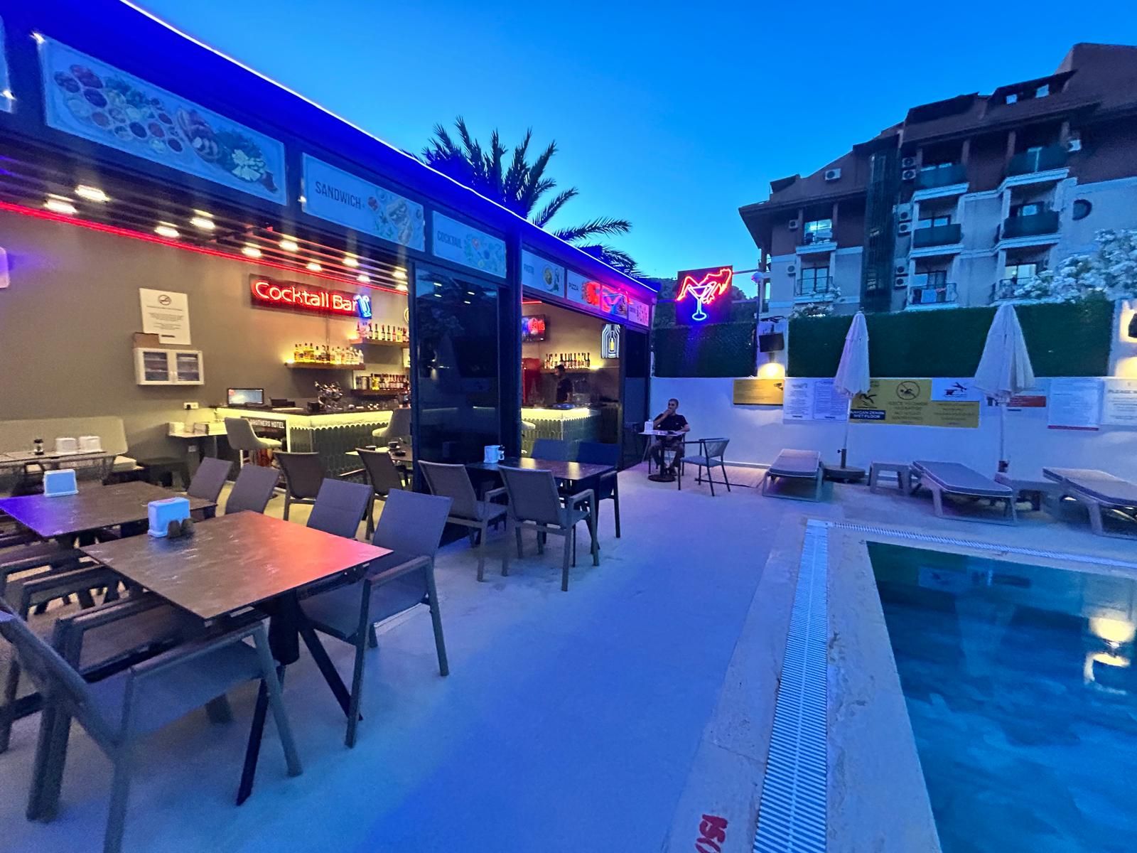 Un área de bar al aire libre con mesas y sillas, una piscina y un edificio con balcones.