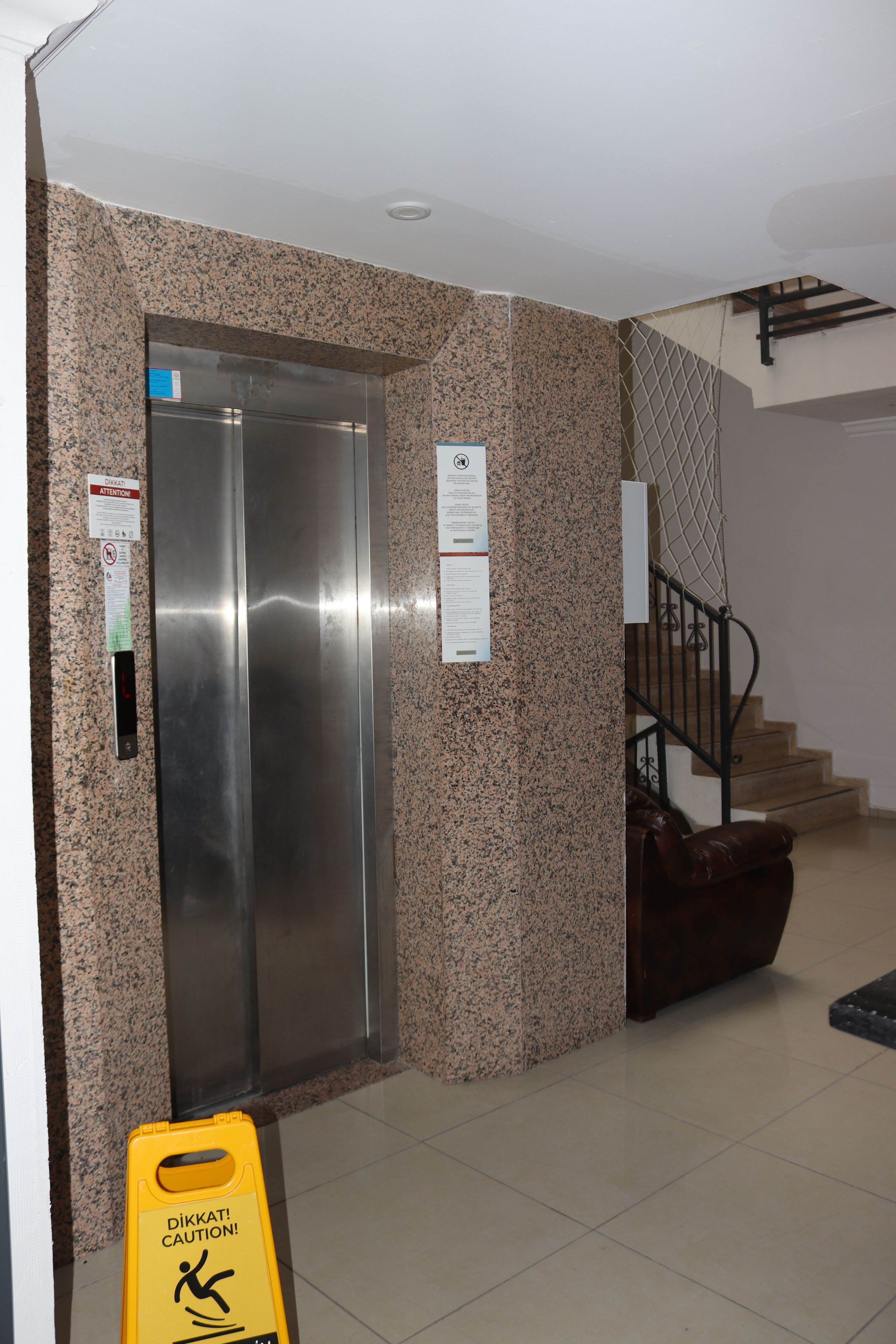 Un ascensor con pantalla digital, un letrero de precaución y una escalera con barandilla de metal.