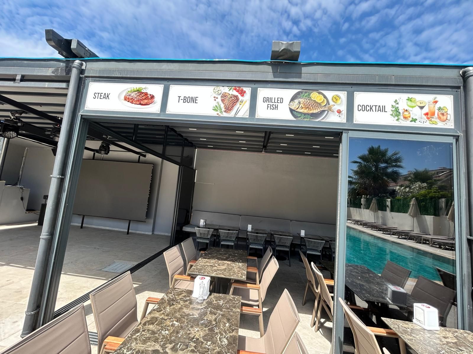 Un área de comedor al aire libre con vista a la piscina, que incluye un menú con opciones de filete, chuletón, pescado a la parrilla y cócteles.