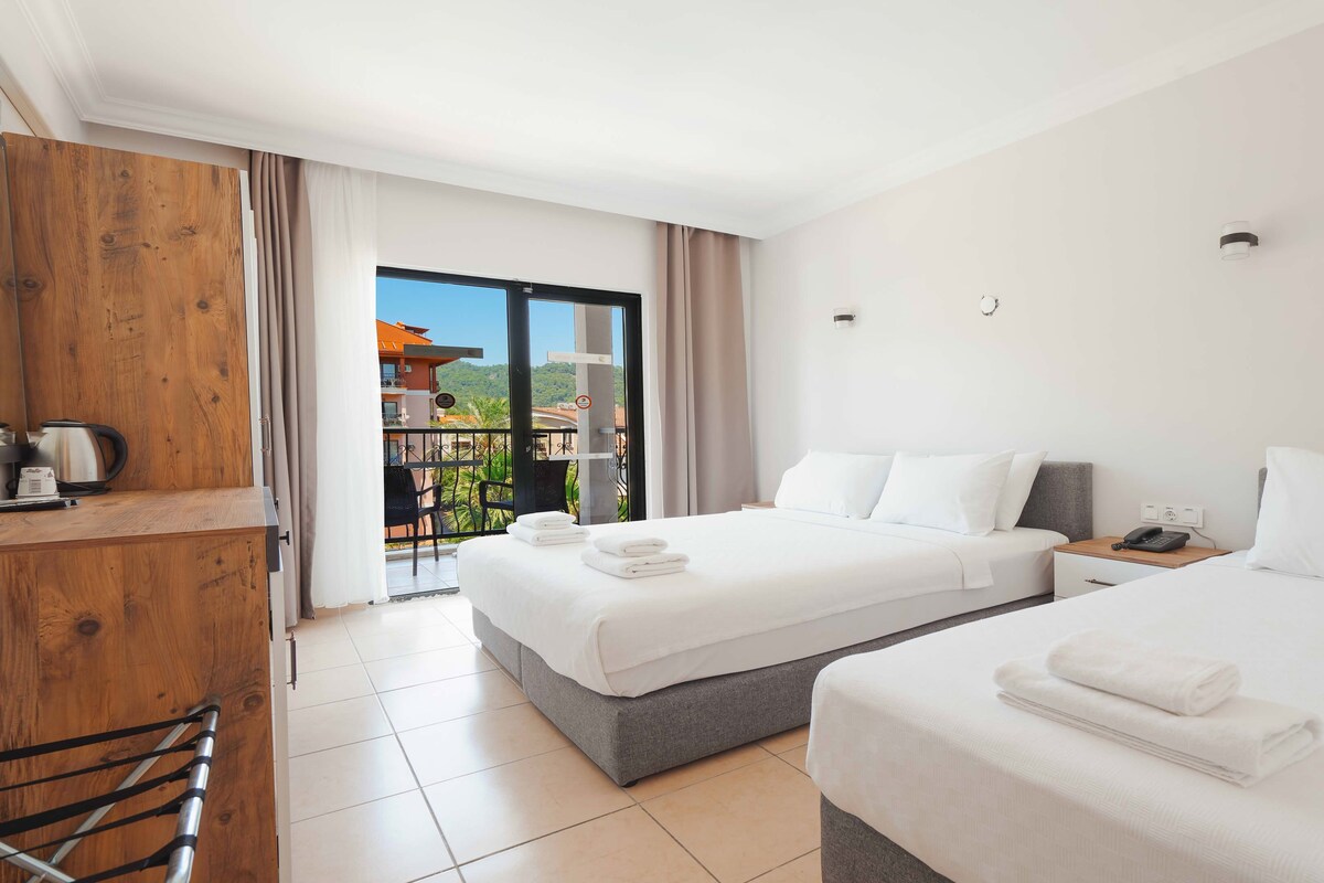 A hotel room with two beds, a desk with a kettle, and a balcony with a view of buildings and greenery.
