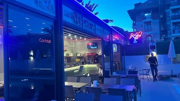 Uma área externa com mesas e cadeiras, um bar com um letreiro de neon e um letreiro neon na fachada do prédio.