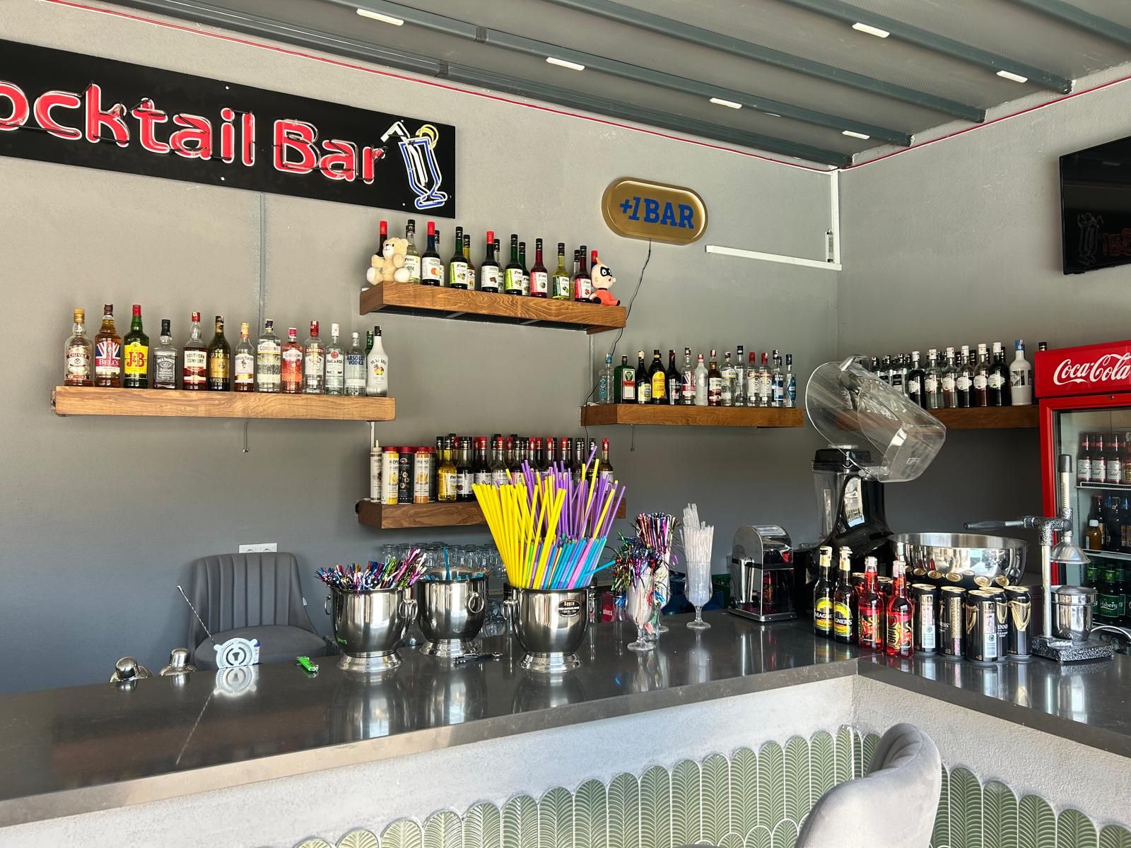 Un bar con estanterías llenas de botellas de licores, un refrigerador de Coca-Cola y un letrero que dice 'Cocktail Bar'.