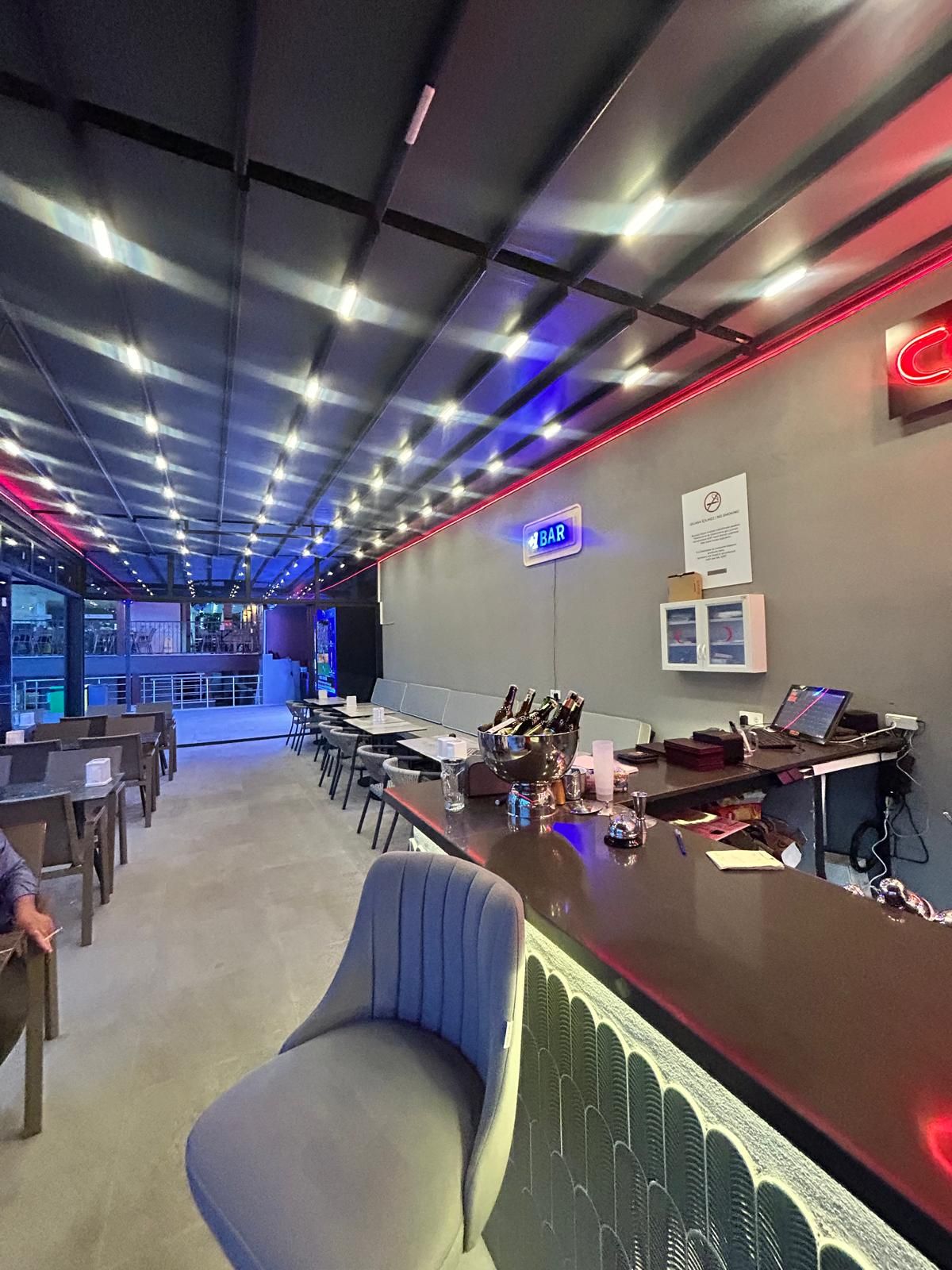 Zona de bar con un letrero de neón, barra y área de estar con mesas y sillas.