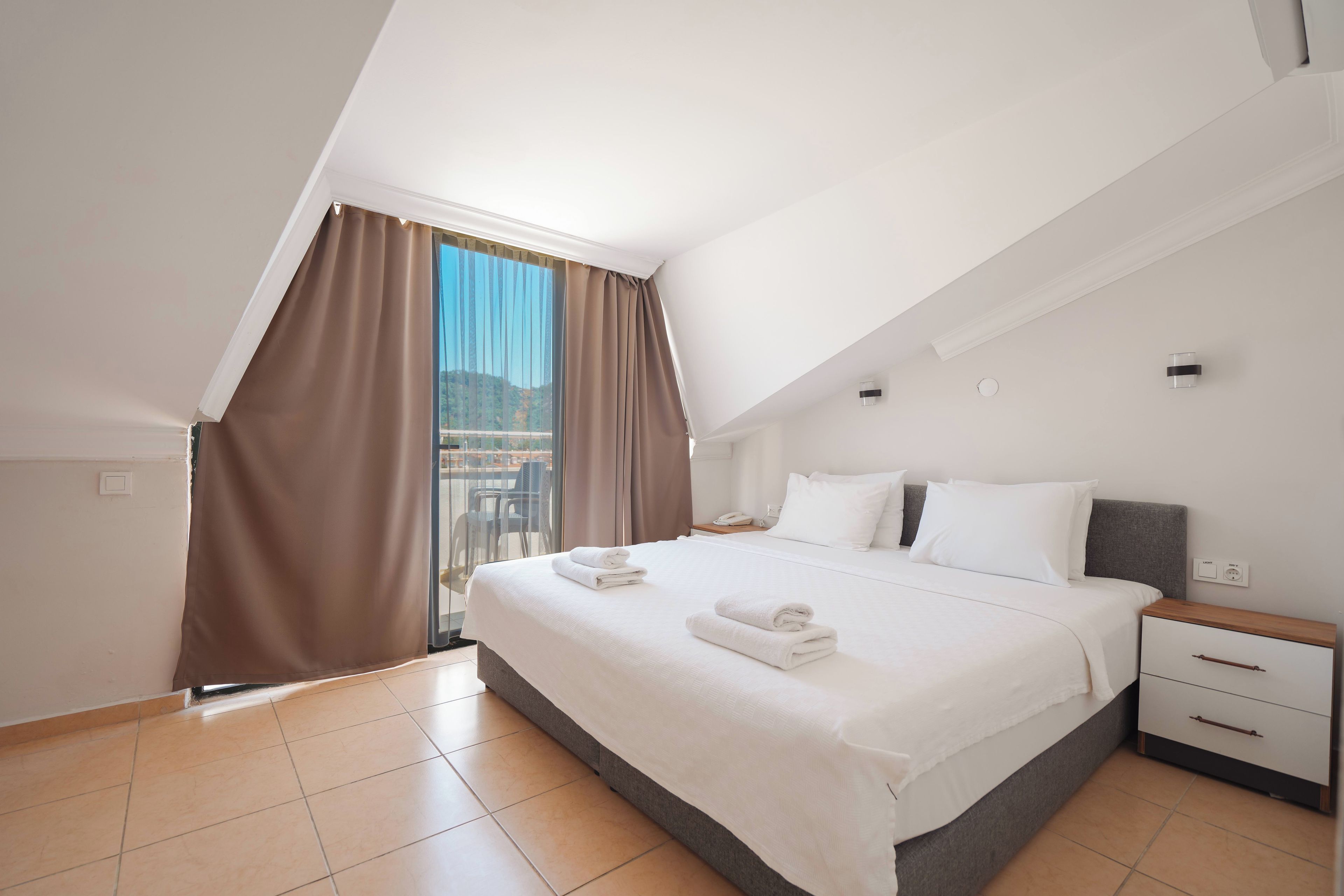 Un dormitorio con cama, mesitas de noche, ventana con cortinas y vistas al exterior.