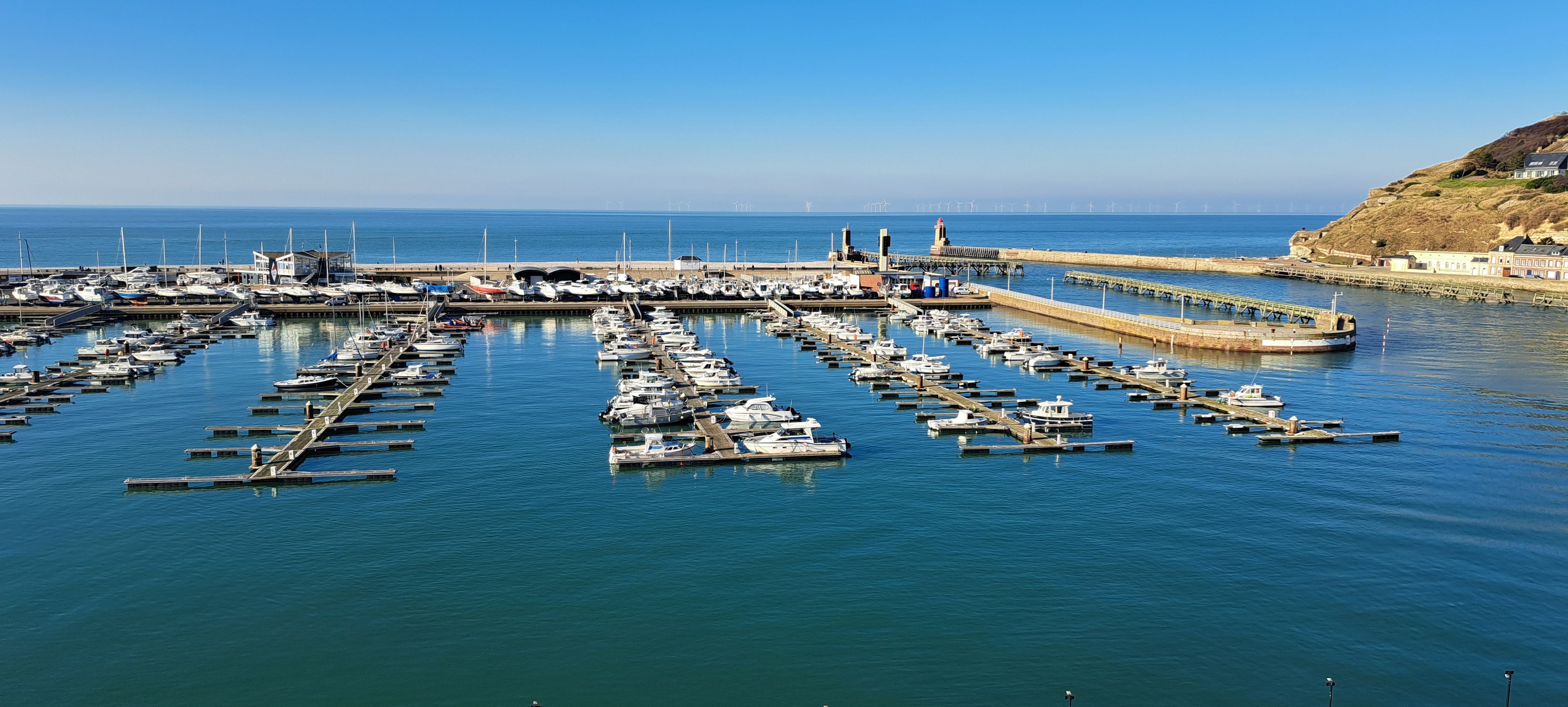 Een jachthaven met talloze boten afgemeerd in kalm water, een steiger die zich uitstrekt over de zee, en een heuvelachtig landschap op de achtergrond.