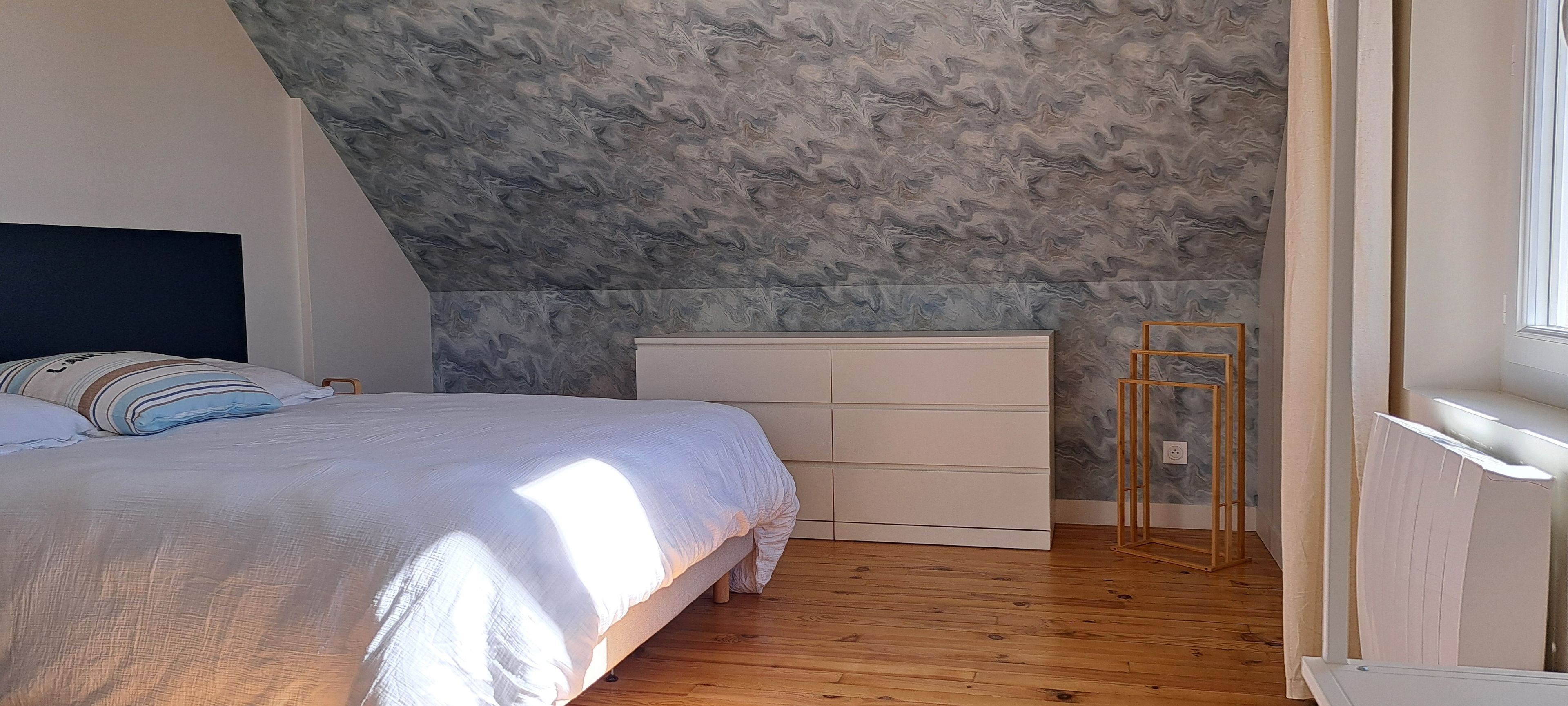 Ein Schlafzimmer mit schrägem Dach, einem Bett mit weißer Bettdecke, Holzboden und einem Fenster auf der rechten Seite.
