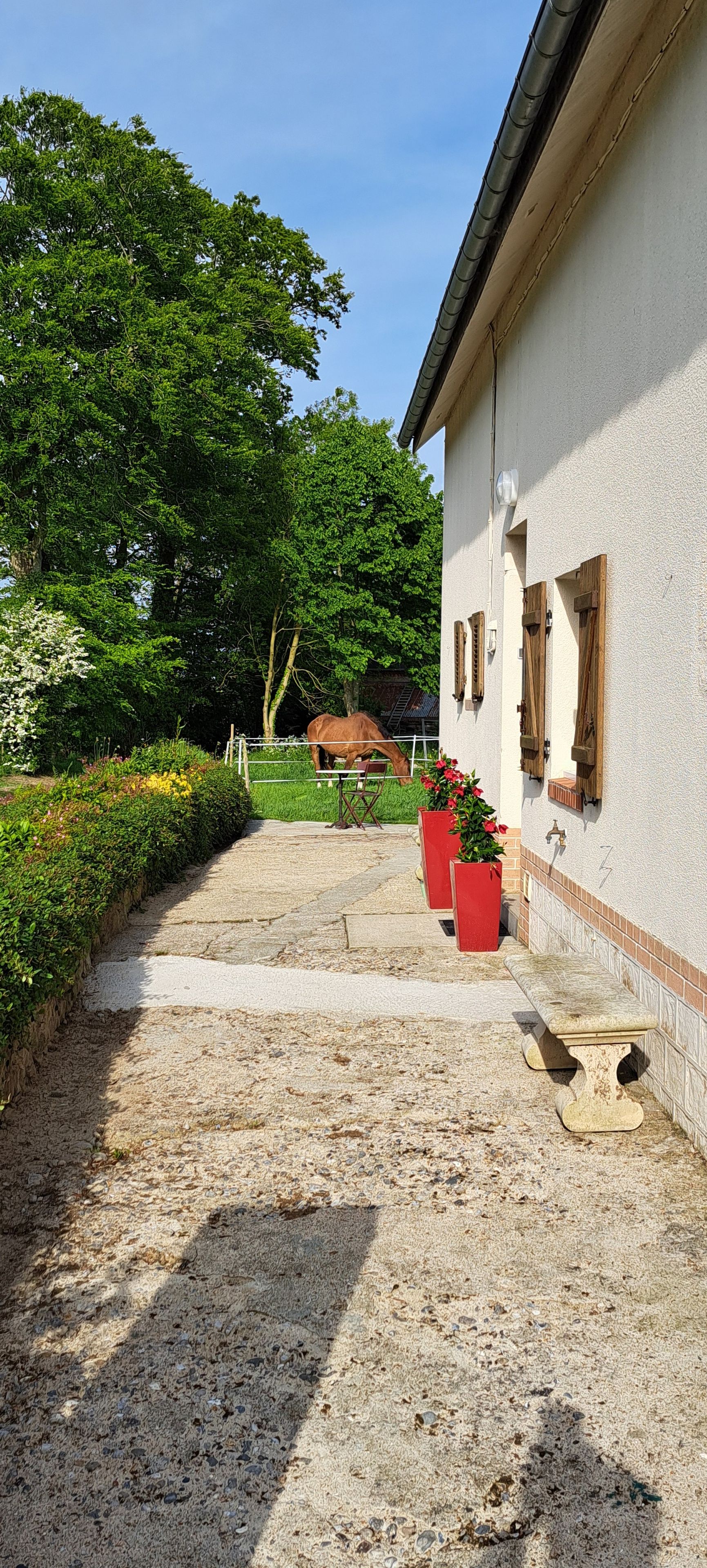 Een gebouw met een grindpad, houten luiken, en een paard dat op de achtergrond graast.