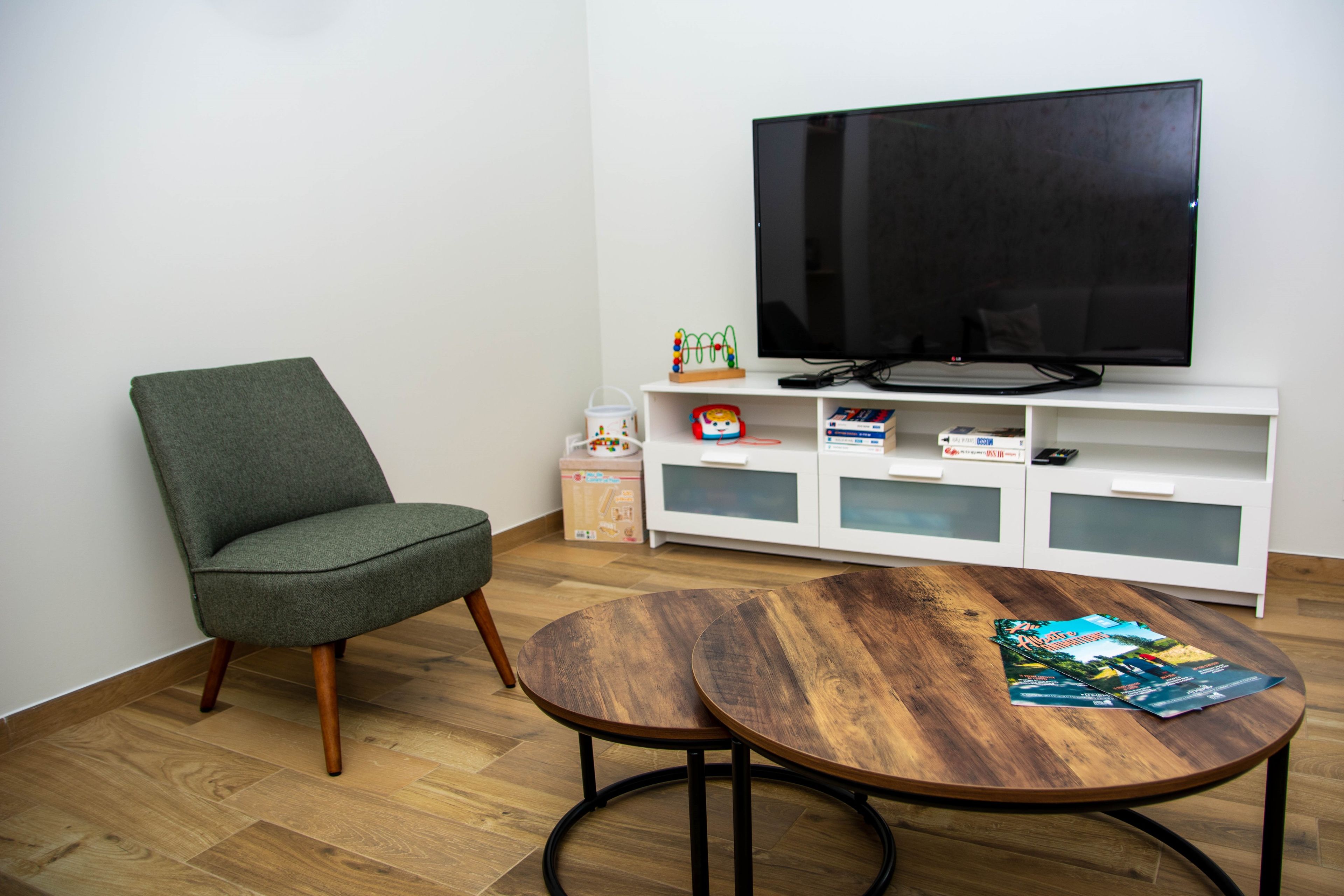 Een woonkamer met een groene fauteuil, een houten salontafel met tijdschriften, een flatscreen televisie op een witte dressoir, en een klein speelgoedje op het meubel.
