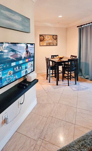 Ein Wohnzimmer mit einem Flachbildfernseher an einem weißen Schrank, einem Esstisch mit vier Stühlen und einem Bild an der Wand.