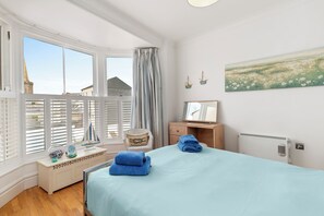 Une chambre à coucher comprenant un lit, une table de chevet, une commode, une fenêtre avec des stores, un tableau et une vue sur les bâtiments à l’extérieur.