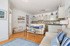 A modern living room with a sofa, a glass coffee table, and a kitchen area with cabinets and a countertop. - 6 Paragon House - St Catherines View - Tenby Sea View (Tenby)