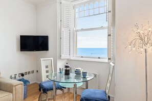 A modern room with a glass table, chairs, a flat-screen TV, a window with shutters, and a decorative plant. - 6 Paragon House - St Catherines View - Tenby Sea View (Tenby)