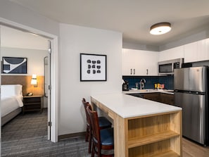 Habitación de hotel con cama, mesita de noche, lámpara, cuadro enmarcado y una kitchenette con microondas, refrigerador y encimera de madera.