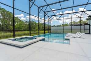 Una piscina amb un banyera d'hidromassatge, envoltada per una zona coberta amb estructura metàl·lica i parets de vidre, amb fons de paisatge de trees i cel blau.