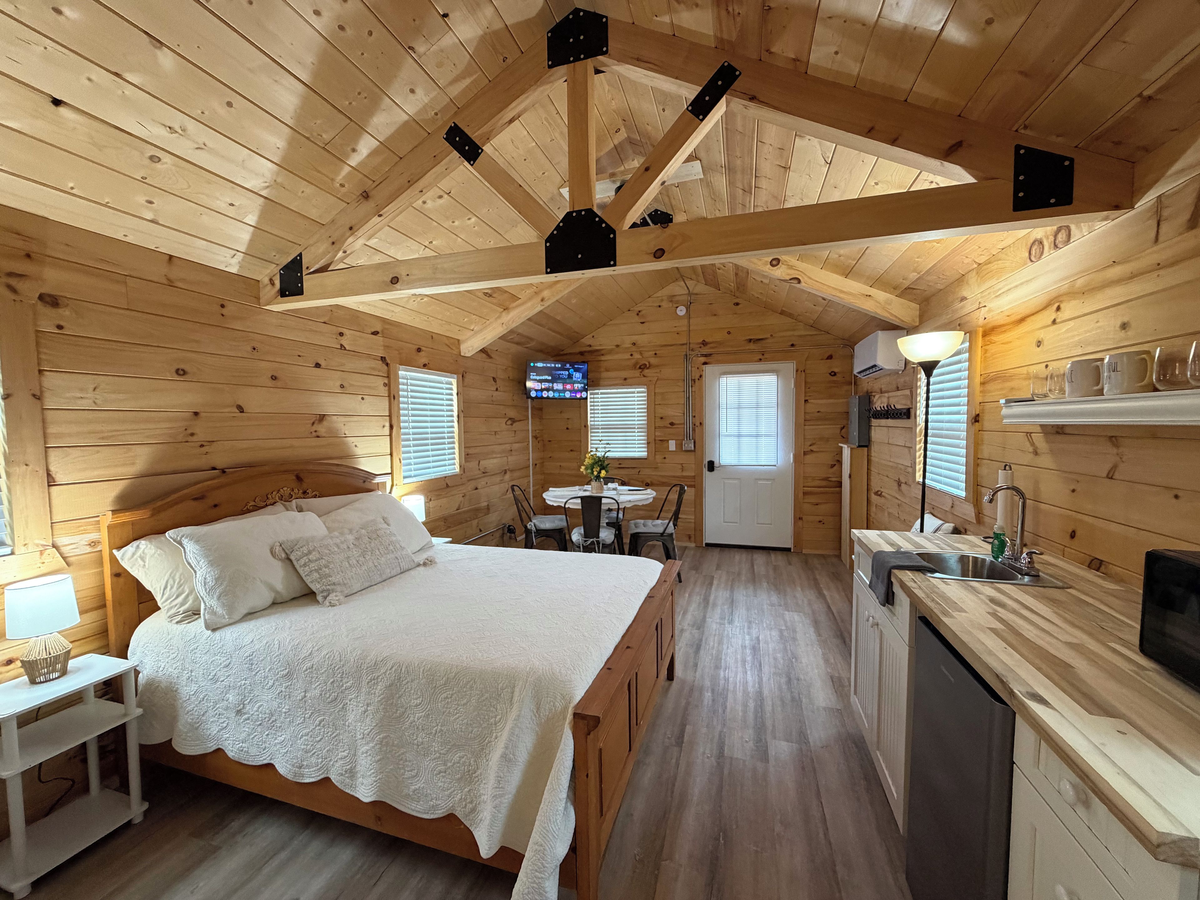 Una cabaña acogedora con interior de madera, una cama con cubrecama blanco, un pequeño comedor y una cocina con fregadero y microondas.