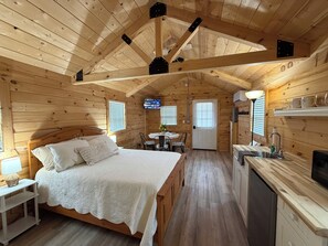 A cozy cabin with a wooden interior, a bed with a white bedspread, a small dining area, and a kitchen area with a sink and microwave.