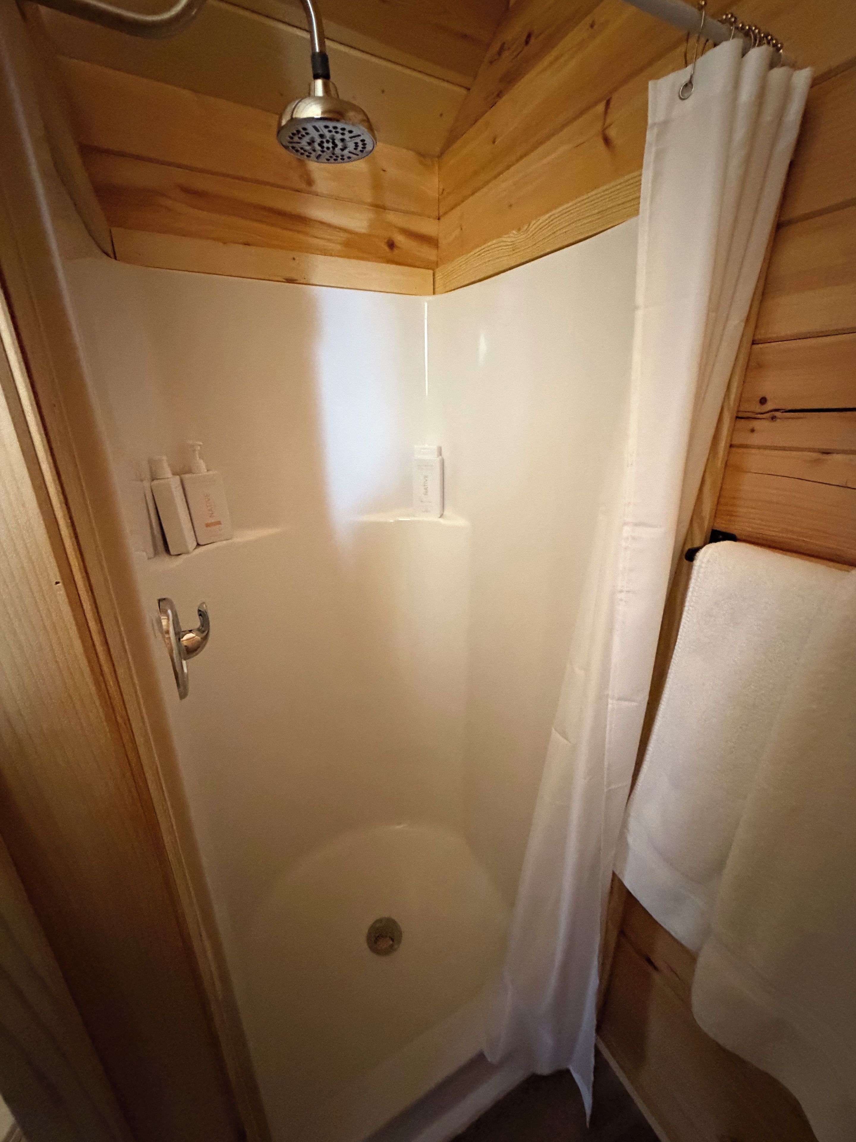 A wooden-paneled shower stall with a showerhead, a white curtain, and a towel hanging on the wall.