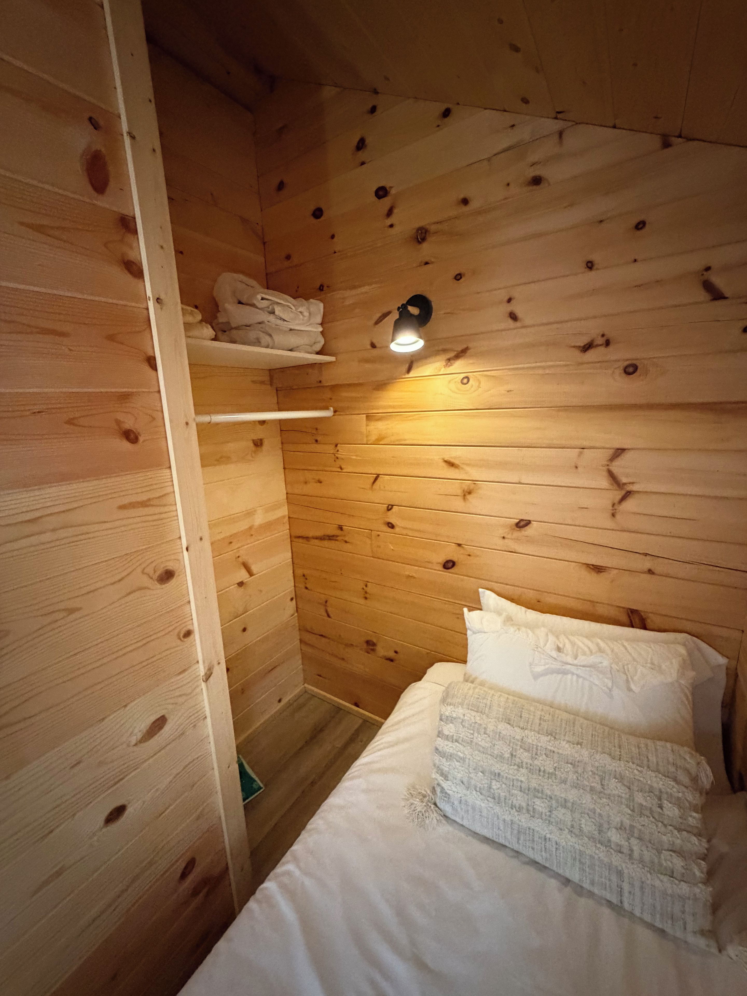 A small, cozy room with wooden walls, a single bed with white bedding, a shelf with folded towels, and a wall-mounted light fixture.