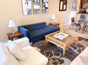 A living room with a blue sofa, a white armchair, a glass coffee table, and a dining area with a table and chairs. - Luxury Las Vegas Strip!  1BR Private Residences Condo ~ 24/7 Pool 2-22 (Las Vegas)