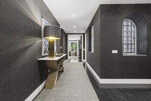 Un couloir avec un tapis à motifs, une lampe fixée au mur et une fenêtre décorative ornée d’un motif géométrique.