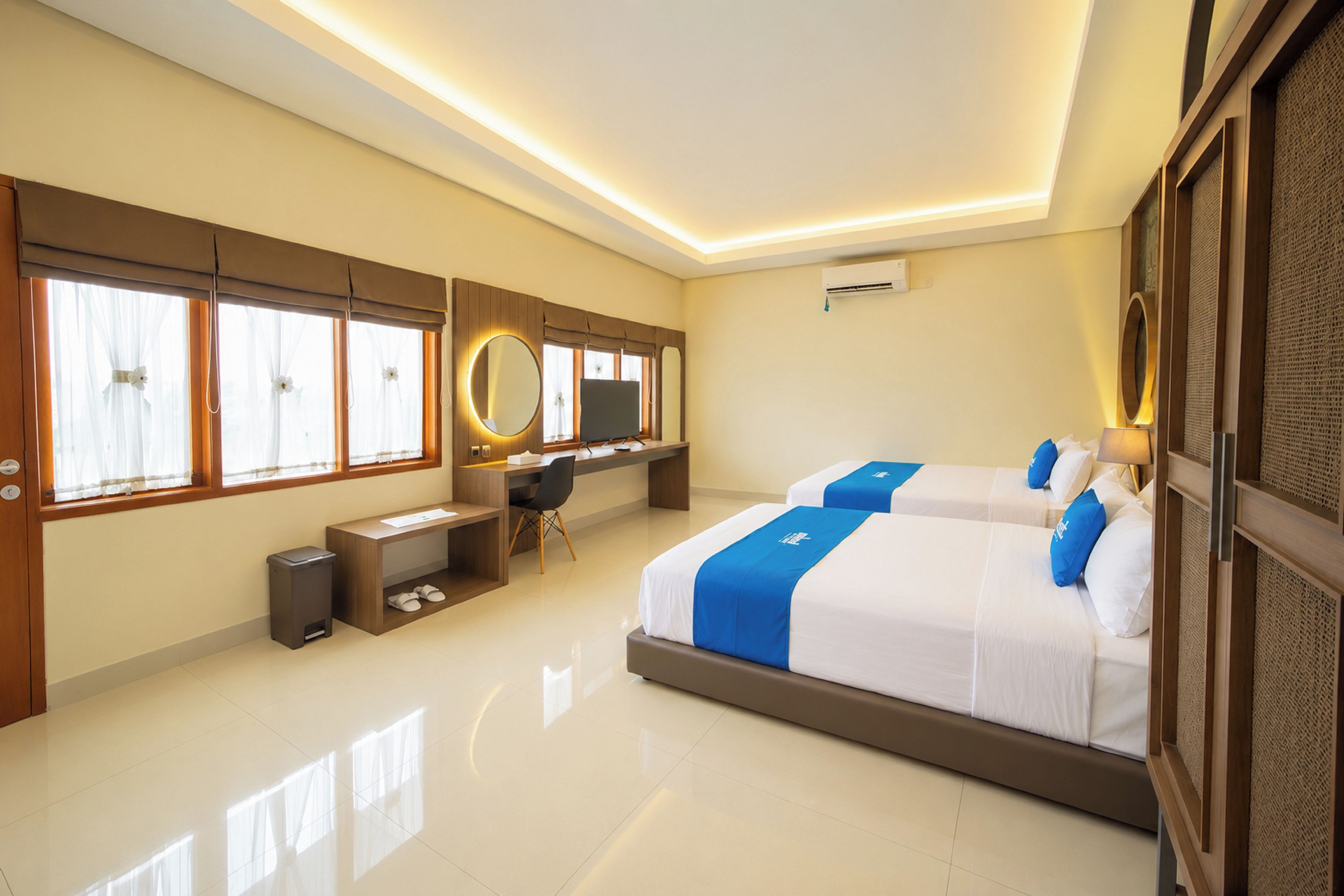 A hotel room with two beds, a desk, a chair, a trash bin, and a large window with curtains.
