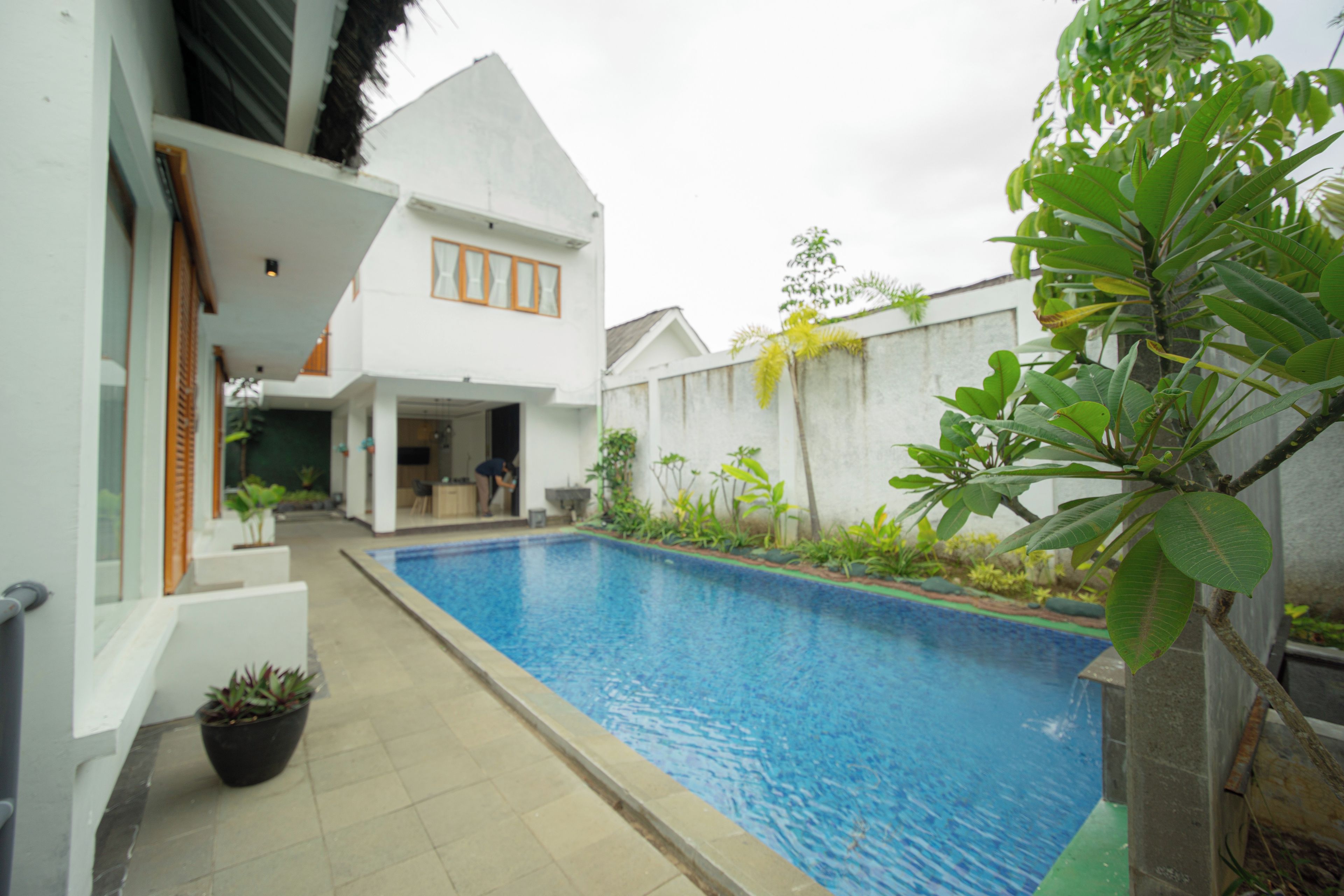 Une piscine avec une terrasse carrelée, un bâtiment blanc doté d’un toit en pente et une grande variété de plantes.