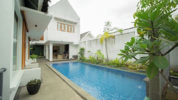 Une piscine avec une terrasse carrelée, un bâtiment blanc doté d’un toit en pente et une grande variété de plantes.