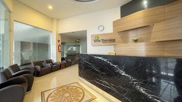 Un hall d’accueil d’hôtel avec un comptoir de réception en marbre noir, des sièges en cuir, une horloge et un panneau indiquant le service de réception.