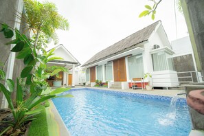 A swimming pool with a tiled edge, surrounded by lush greenery and a white building with a thatched roof.