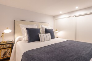 A neatly made bed with a dark grey blanket, white linens, and several pillows in shades of white and dark blue. There are two bedside lamps with gold bases, and a nightstand with a decorative bird painting. - Madrid Duplex | Barrio de las Letras (Madrid)