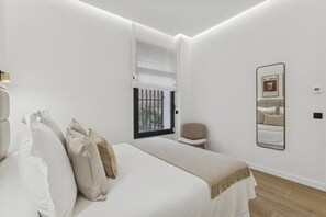 A bedroom with a bed, pillows, a chair, a window with blinds, and a wall-mounted mirror. - Spacious Home in the Heart of Chamberí (Madrid)