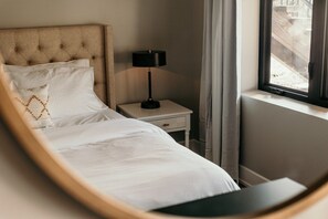 Ein ordentlich bezogenes Bett mit weißen Leinen, ein Nachttisch mit Lampe und ein Fenster mit Vorhängen.