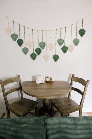 A round wooden table with two chairs, a small white object on the table, and a wall hanging with heart-shaped decorations.