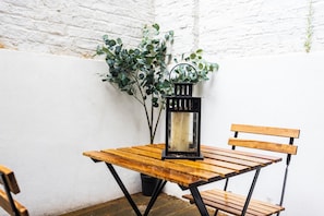Una mesa de madera con un farol negro, un banco y una planta en maceta, todo frente a una pared de ladrillo blanco.