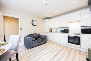 A modern kitchen with white cabinets, a built-in oven, and a dining area with a grey sofa and a white chair. - Comfy & Spacious|2 Bedroom Apartment|Seconds from Hammersmith Central Station (London)