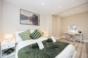 A hotel room with a bed, a desk, a chair, a lamp, and a painting on the wall. - Comfy & Spacious|2 Bedroom Apartment|Seconds from Hammersmith Central Station (London)