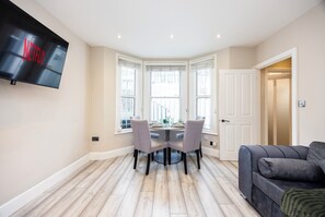 A living room with a flat-screen TV mounted on the wall, a dining area with a table and chairs, and a grey sofa. - Comfy & Spacious|2 Bedroom Apartment|Seconds from Hammersmith Central Station (London)
