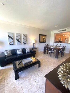 A living room with a black sofa, a coffee table, and a dining area with a table and chairs. There are abstract wall art pieces and a mirror above the dining table. - Luxury Las Vegas Strip!  2BR Condo ~ Sphere view ~ 24/7 Pool 3-24 (Las Vegas)