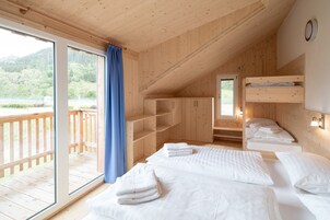Una habitación con paredes de madera y un balcón, que incluye dos camas con ropa blanca y toallas, además de un ventanal con cortina.