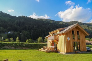 A wooden cabin with a balcony, situated in a grassy field with a forested mountain in the background. - 002 MW5 in Murau (Murau)