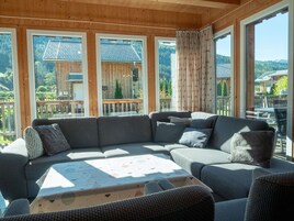Habitación con paneles de madera, un sofá seccional grande, una mesa y vistas a una terraza de madera y edificios.