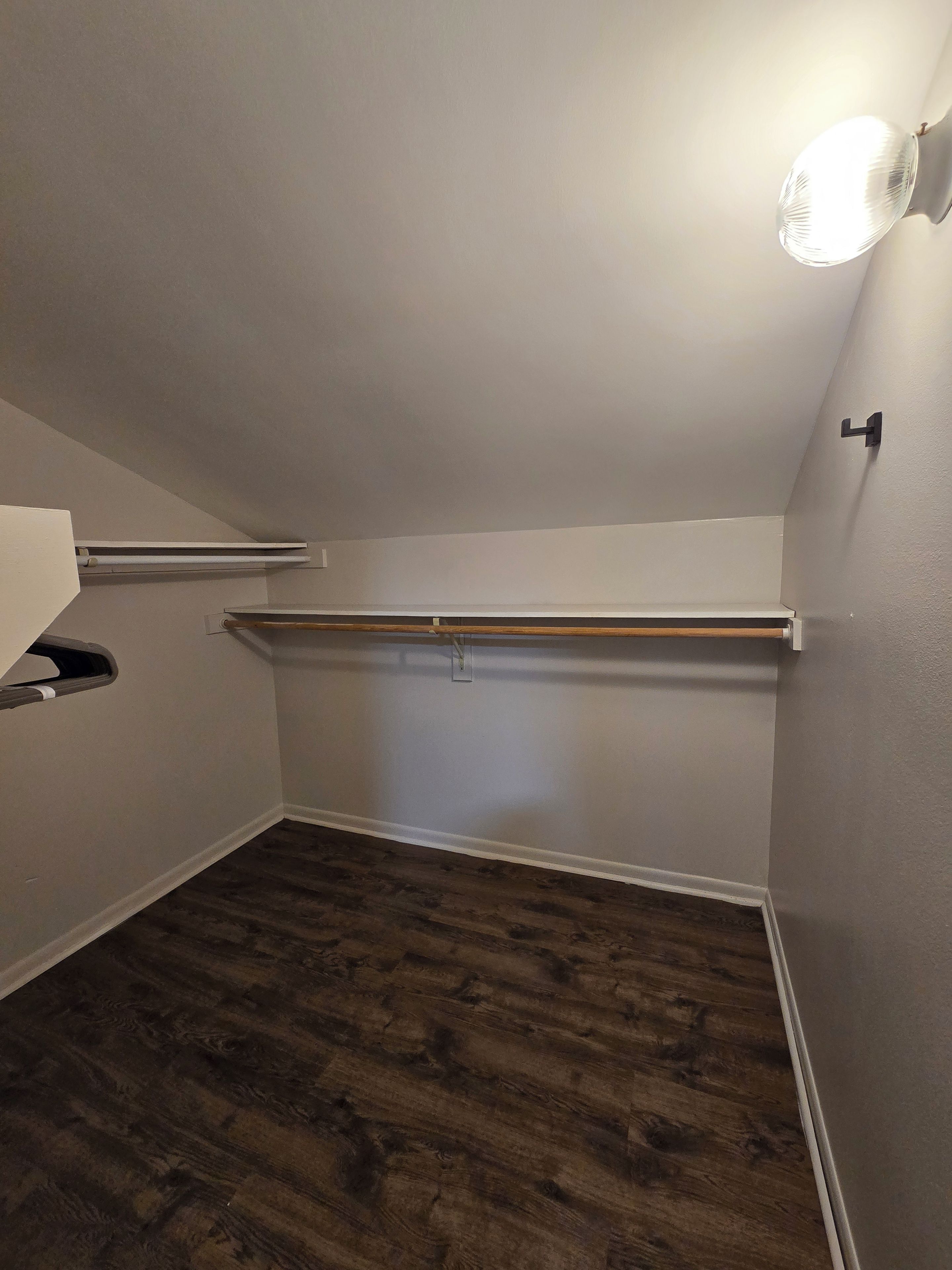 Una stanza piccola, scarsamente illuminata, con soffitto spiovente, pavimento in legno e una singola lampada.