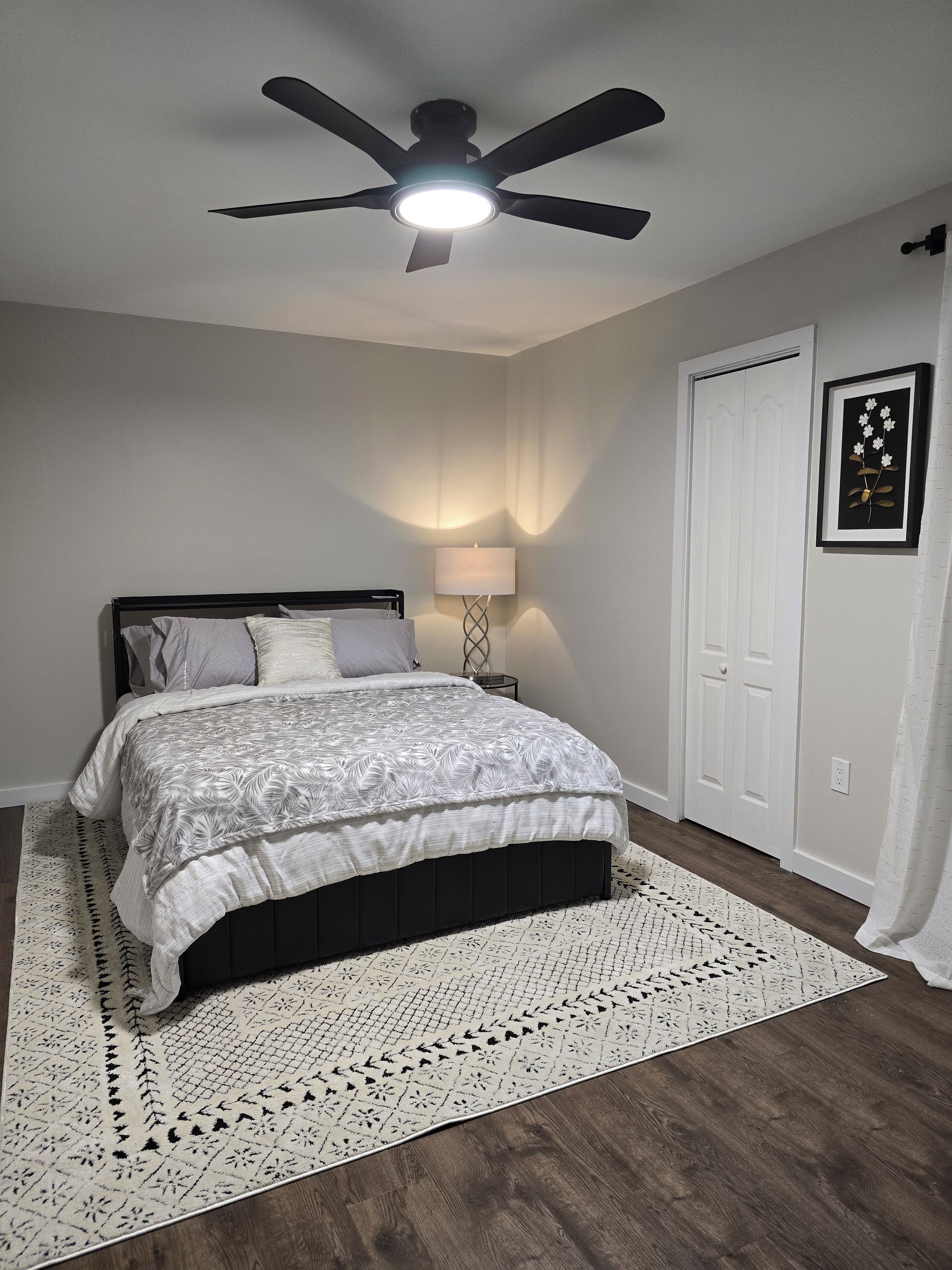 Un dormitorio con cama, ventilador de techo, lámpara de noche, puerta y un cuadro en la pared.