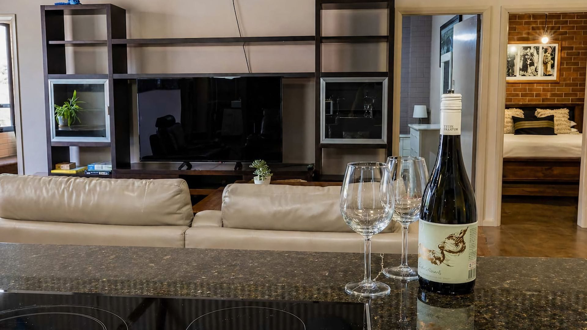 A modern living room with a sofa, a flat-screen TV, a wine bottle, and glasses on a counter.