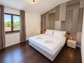 Ein modernes Hotelzimmer mit einem großen Bett, einem Nachttisch, einem Fenster mit Vorhängen und Blick auf Grünflächen.
