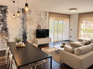 Een moderne woonkamer met een eethoek, een flatscreen televisie en een groot raam met gordijnen.