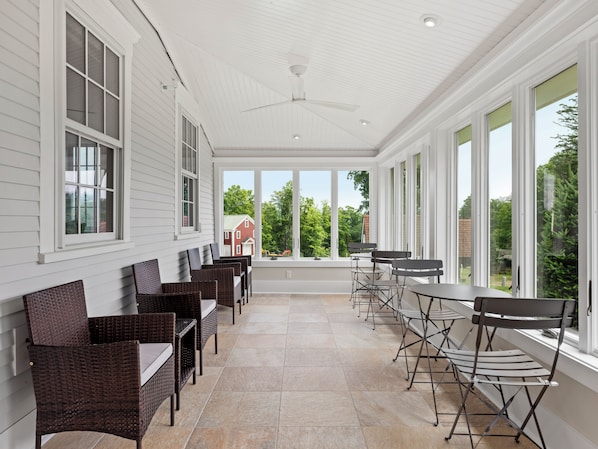 A covered patio with wicker furniture and large windows offering views of a red building and greenery.