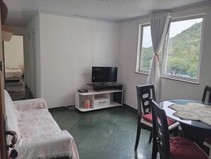 A room with a bed, a TV on a stand, a dining table with chairs, and a window with curtains.