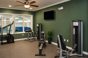 Ein Fitnessraum mit Trainingsgeräten, einem Deckenventilator, einem Fernseher und einem Fenster mit Blick auf einen Innenpool.